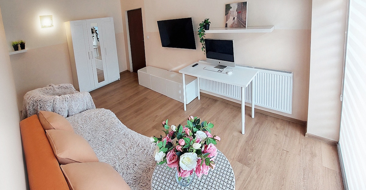 A modern living area is shown with light wooden flooring and soft beige walls. A comfortable sofa is arranged in front of a white low-profile cabinet and two flat-screen TVs. A stylish table adorned with a bouquet of flowers adds a touch of color to the space.