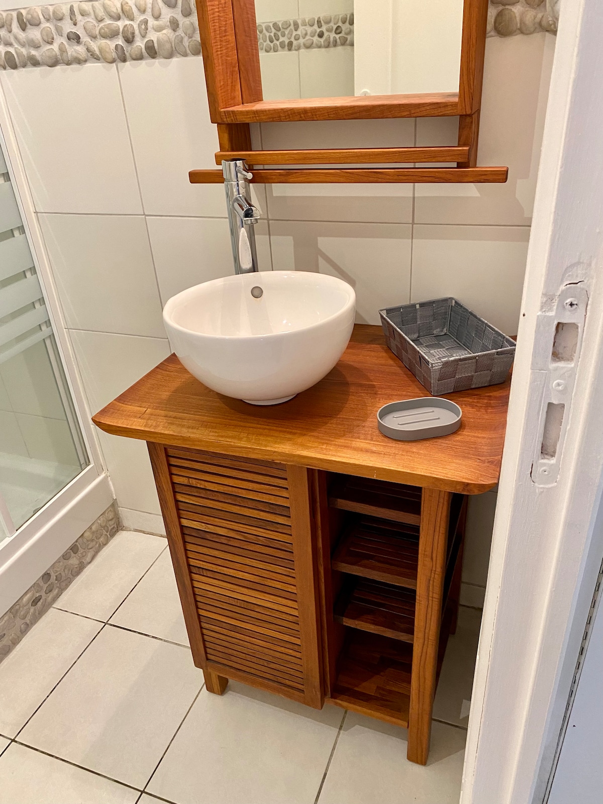 A modern bathroom sink area features a round, white vessel sink atop a wooden vanity with slatted doors, providing storage below. A rectangular mirror is mounted above, and a small decorative basket sits beside a soap dish. The floor is tiled in neutral tones.