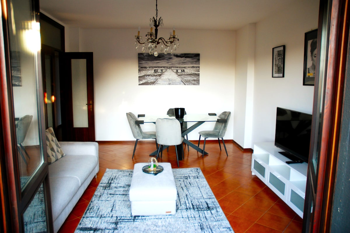 The living area features a modern, light-filled space with a glass dining table and four upholstered chairs. A spacious sofa is positioned adjacent to a sleek entertainment unit containing a television. A decorative rug and an eye-catching chandelier add to the inviting decor.