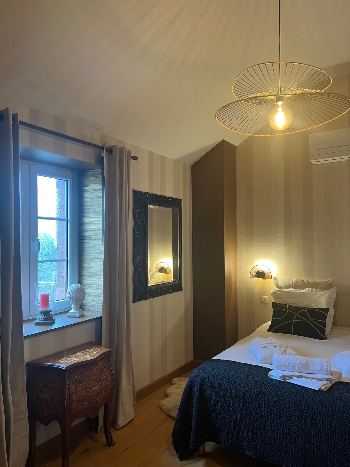 A cozy bedroom features a spacious bed with crisp linens and a decorative throw. Natural light enters through a window adorned with sheer curtains, framing a glimpse of the outdoors. An ornate mirror reflects soft lighting from an elegant ceiling fixture and a nearby wall sconce.