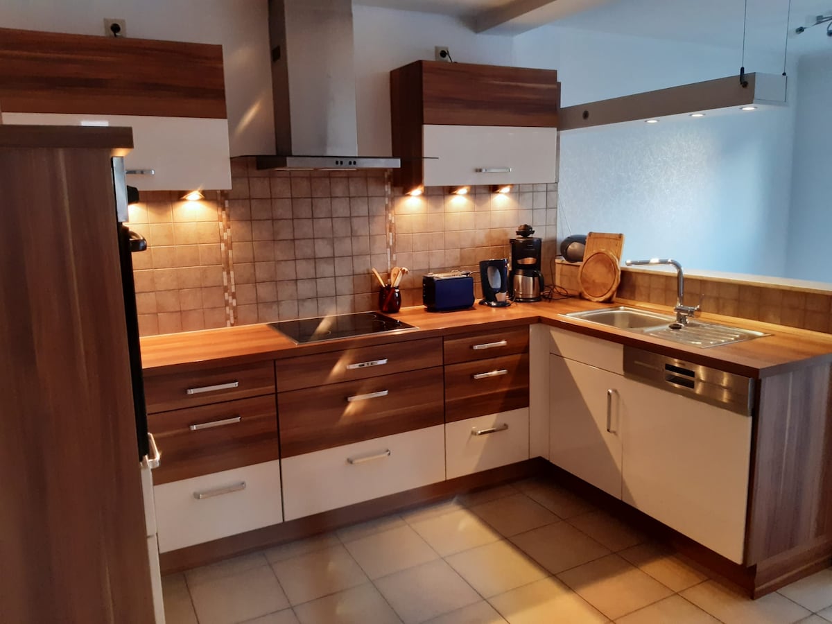 A modern kitchen is equipped with sleek wooden cabinetry and contemporary appliances. Soft lighting highlights the countertop, which features a sink and an induction cooktop. The space is designed for functionality, with a coffee maker and kitchen essentials visible.