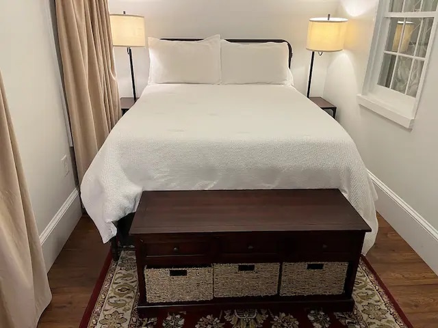 A spacious bed is dressed in crisp white linens, flanked by two bedside lamps that provide soft lighting. A wooden bench with storage baskets is positioned at the foot of the bed, situated on a patterned area rug that complements the warm wooden floor.