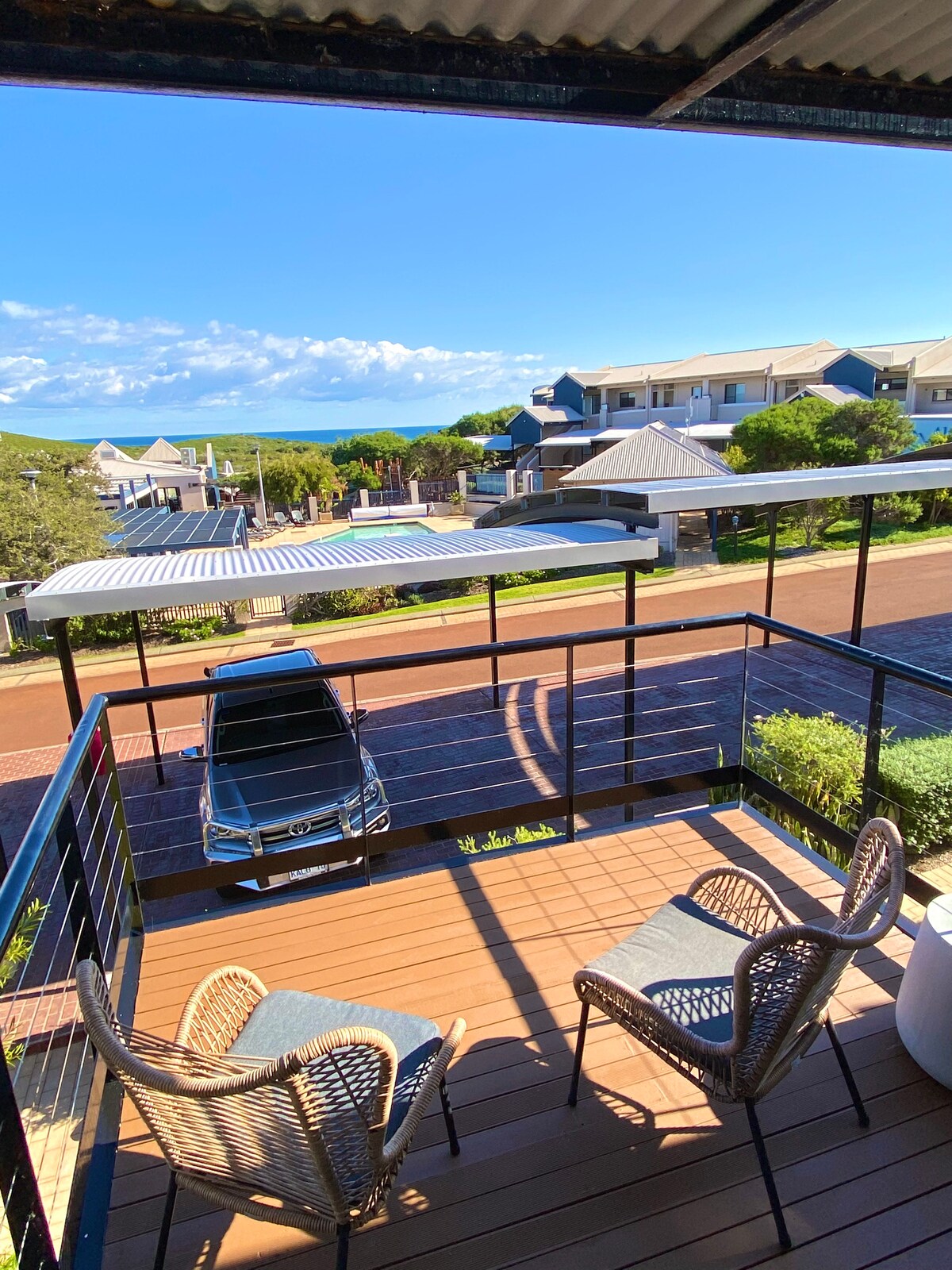 A balcony features two stylish chairs positioned for coastal views, overlooking a neighborhood and the ocean in the distance. The deck is lined with greenery, and sunlight casts soft shadows across the wooden surface.