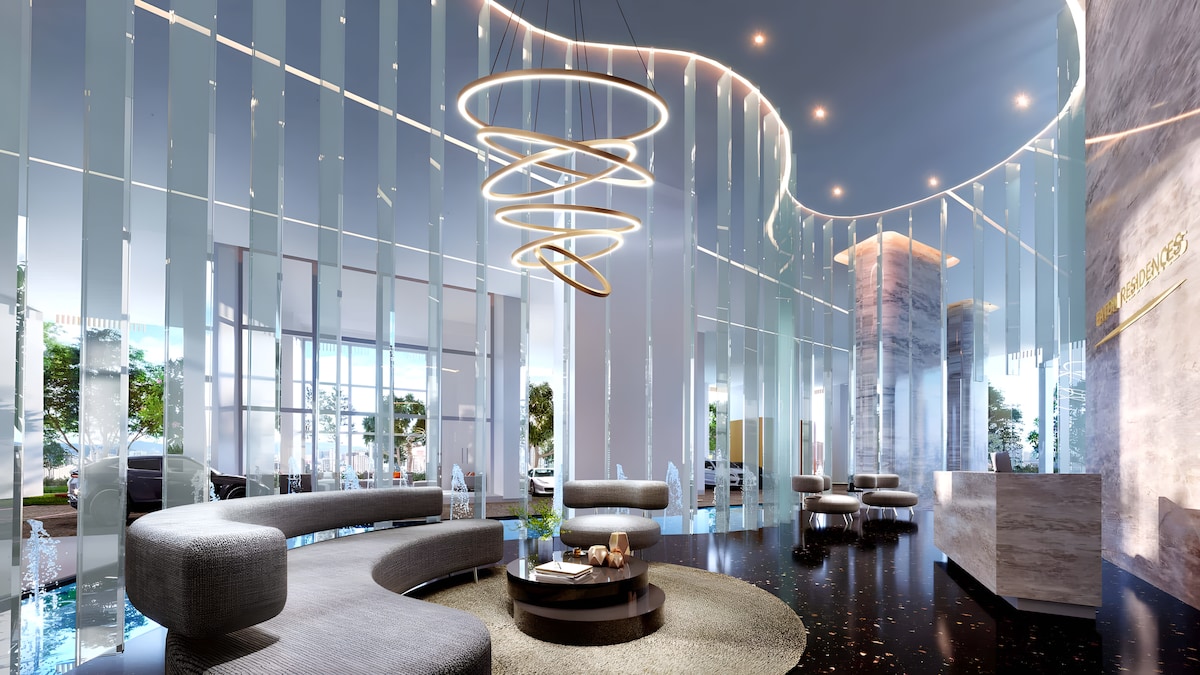 The lobby area is characterized by sleek, modern design elements. Curved seating arrangements are complemented by a unique, spiral light fixture hanging overhead. The space is brightened by natural light filtering through large windows, with greenery visible in the background, creating a refreshing ambiance.