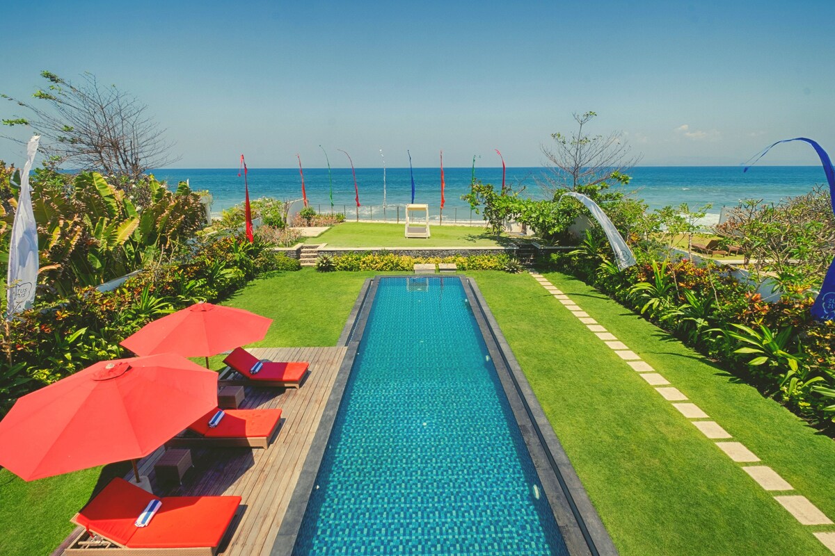 An outdoor area features a long, turquoise swimming pool surrounded by lush greenery. Two bright red umbrellas provide shade over lounge chairs placed on a wooden deck. The view extends towards the tranquil ocean, showcasing a clear blue sky and gentle waves.