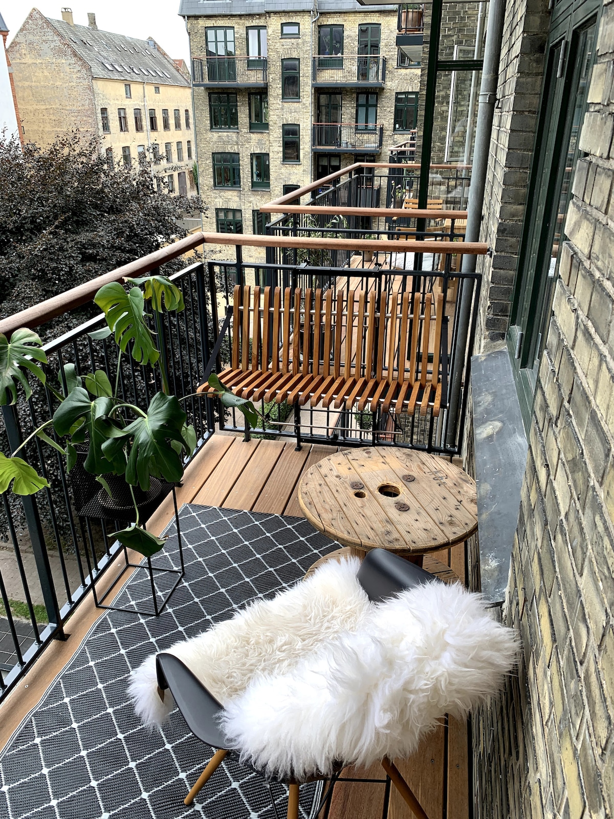 A cozy balcony space features a wooden bench swing and a round table. A plush, white faux fur throw adds comfort to a sleek chair. Green foliage is positioned nearby, and a patterned rug enhances the area. The backdrop includes charming building facades.