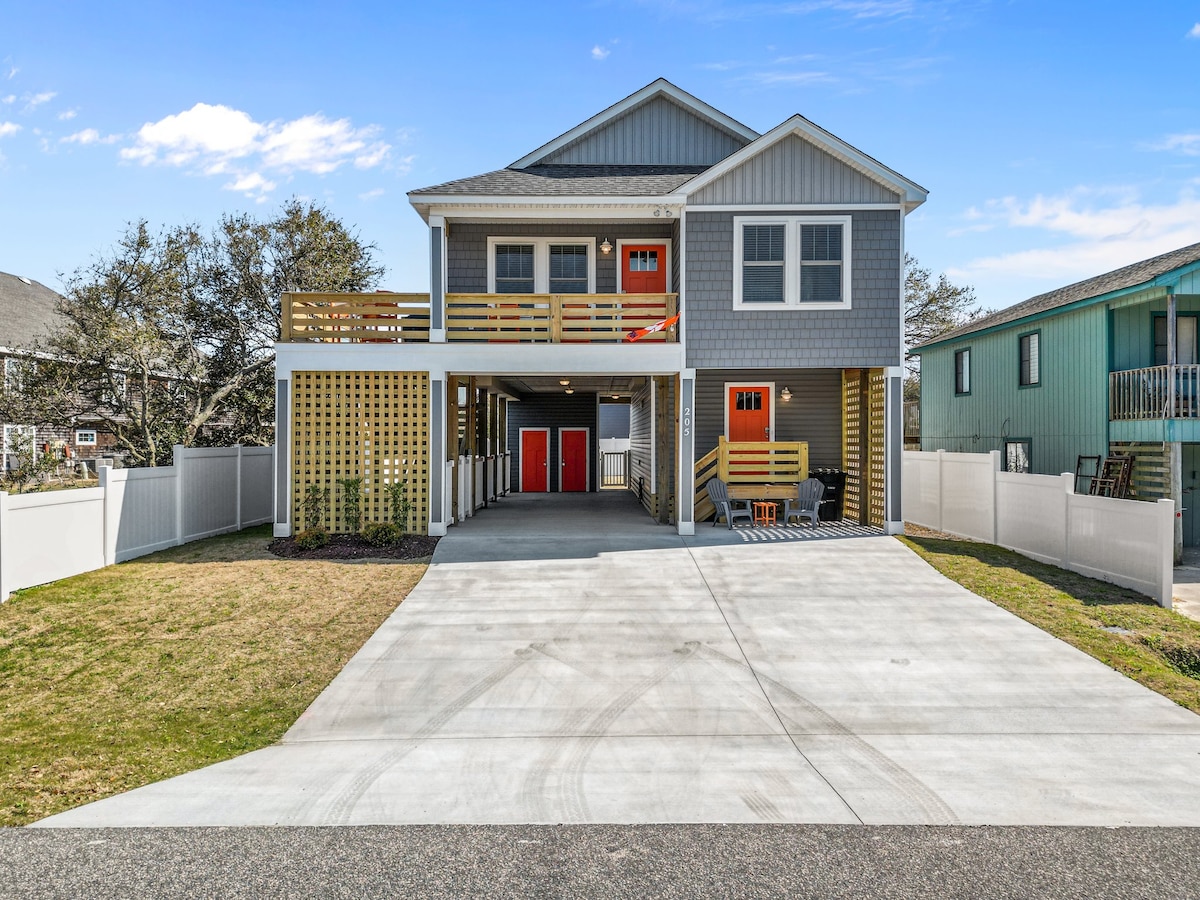 The exterior of a two-story home features a grey façade with yellow accents and red doors. A spacious driveway leads to a covered carport. Outdoor seating is visible under the deck, while a well-kept lawn and white fencing frame the property.