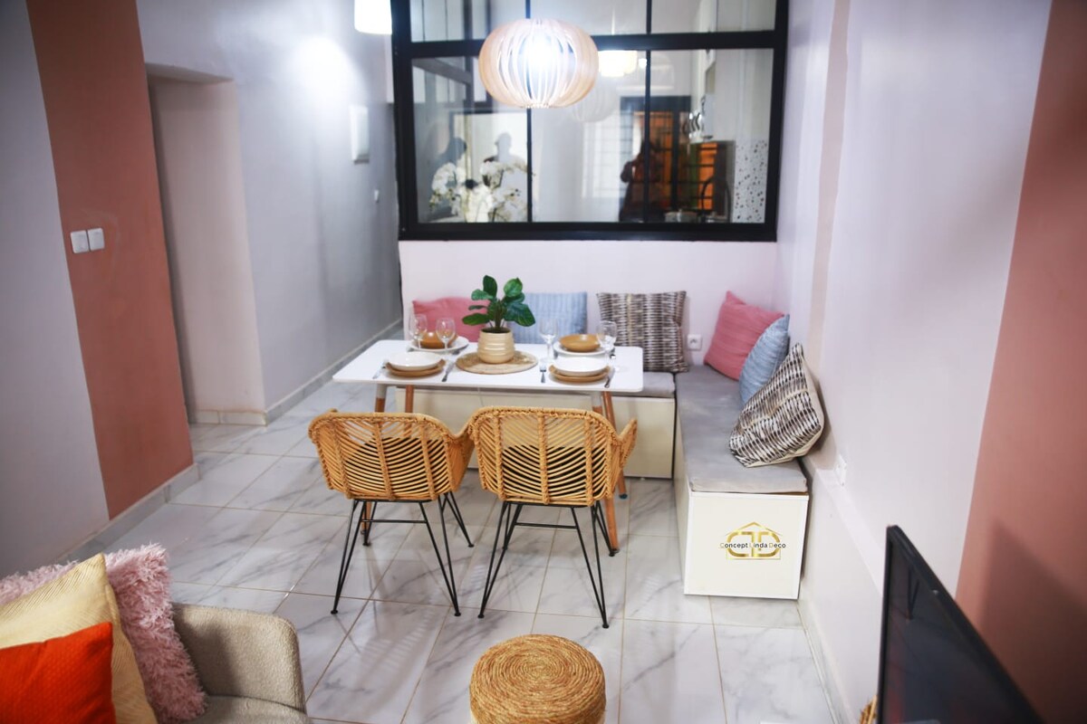 The dining area features a light wooden table surrounded by four woven chairs. A decorative plant is placed at the center of the table, with neatly arranged dinnerware. Soft cushions adorn a nearby seating area, enhancing the overall cozy ambiance of the space.