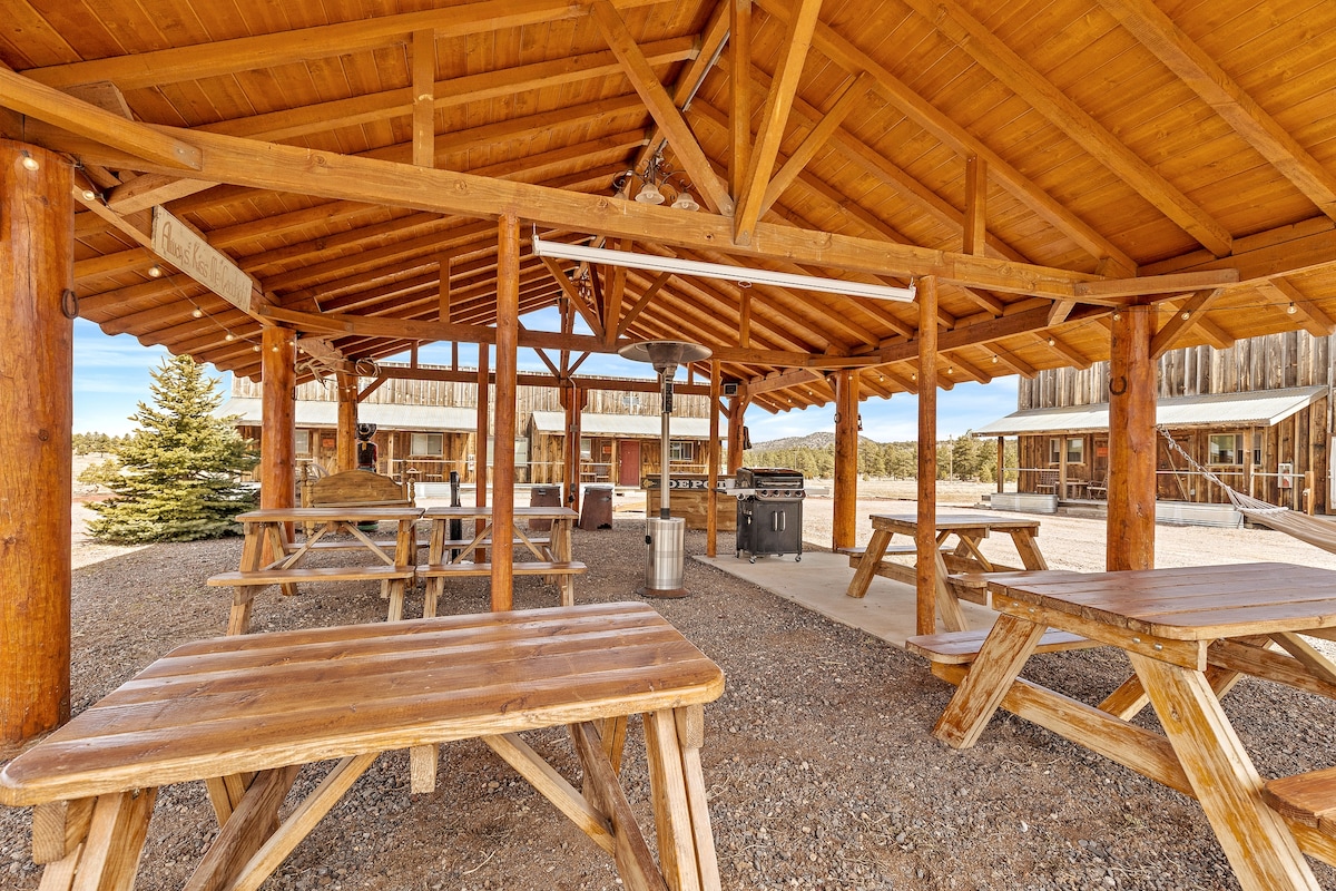 An open pavilion features wooden picnic tables oriented for gatherings, with a grill positioned nearby. The structure is supported by sturdy wooden beams, providing shade and a rustic charm. Gravel flooring surrounds the area, creating a natural setting amidst the mountains.