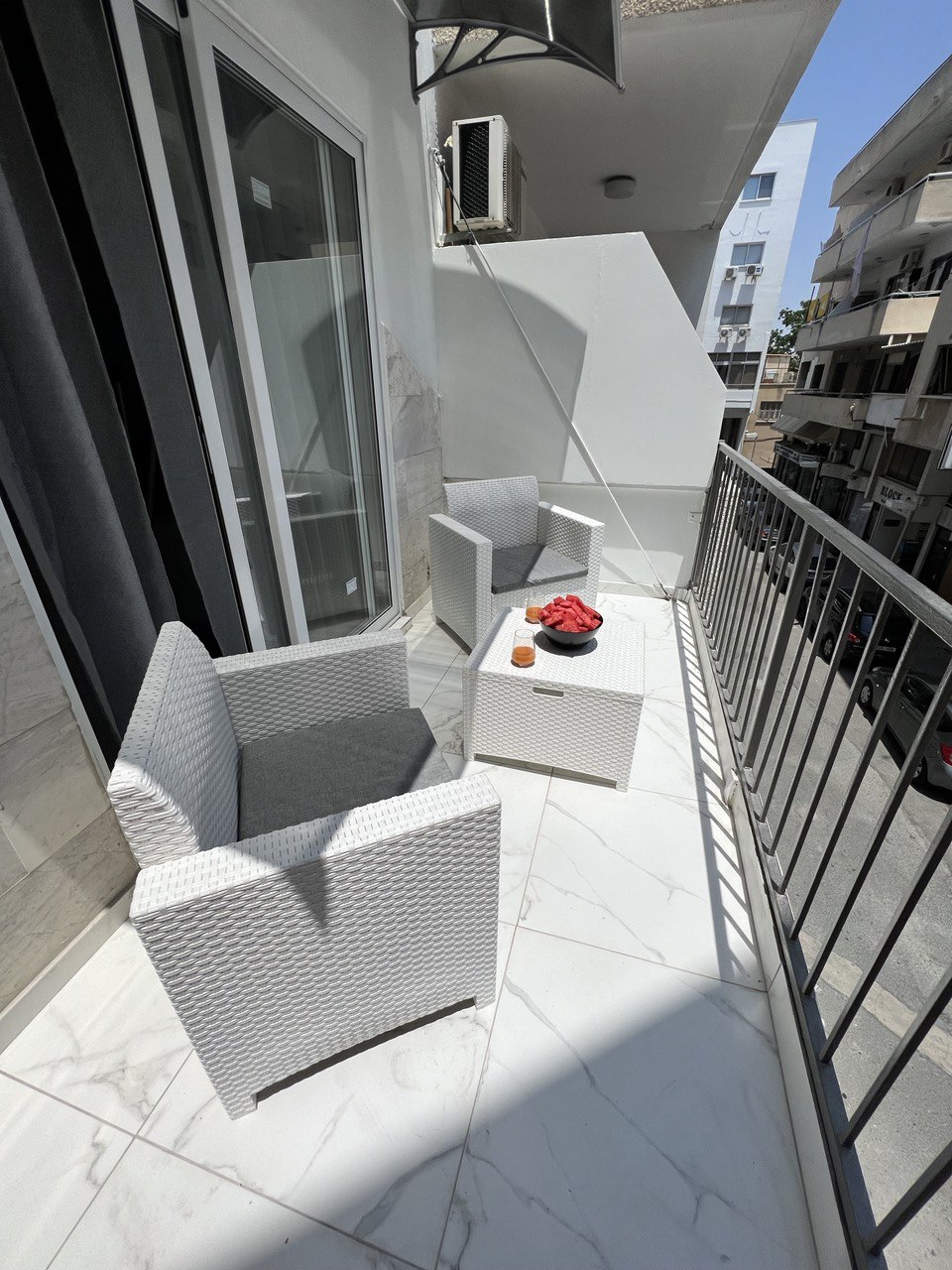A balcony area is presented with two textured armchairs and a small table. A bowl of fruit sits on the table, while the surrounding space features a smooth, tiled floor and an adjacent building view.