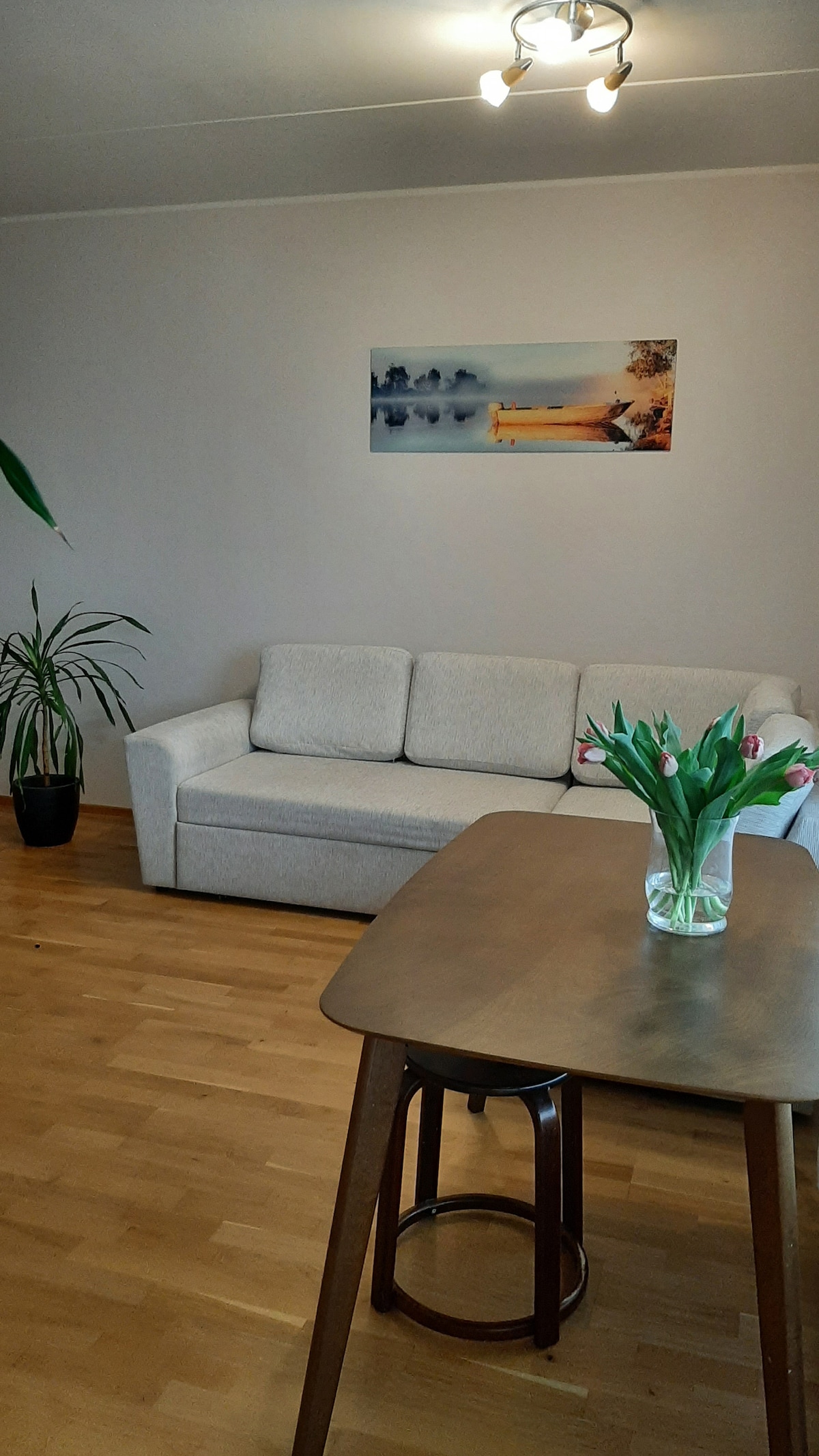 A light and airy living space features a soft sofa against a neutral wall. A dining table with a stool is prominently displayed, accompanied by a vase of fresh tulips. A potted plant adds a touch of greenery to the room.