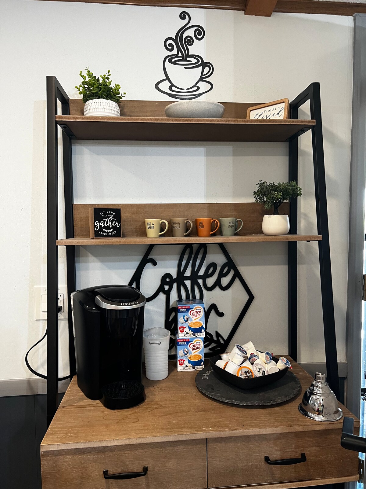 A coffee station is presented with a modern black and wood shelving unit. Ceramic mugs in various colors are neatly arranged on two shelves above a single-serve coffee maker. Creamer and sugar packets are displayed on a round tray, adding convenience for guests.