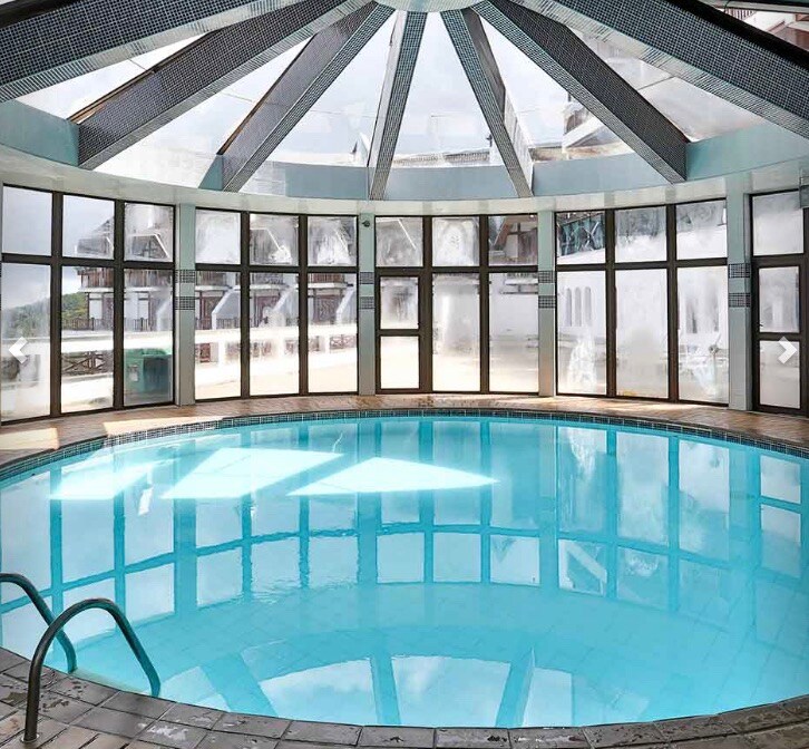A circular indoor pool is surrounded by large glass windows, allowing natural light to illuminate the space. The pool's clear water reflects the shape of the ceiling, which features a geometric design. Steps lead into the pool, enhancing accessibility.
