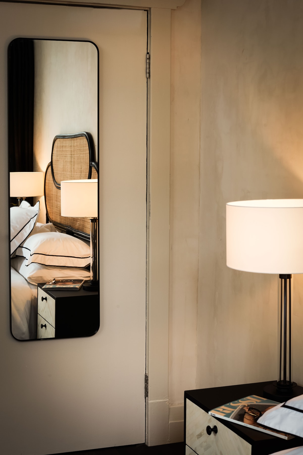 A full-length mirror is mounted on a light-colored door, reflecting a cozy sleeping area with a plush bed and bedside lamp. The lamp casts a warm glow, while a magazine rests on the nightstand, adding to the serene ambiance.