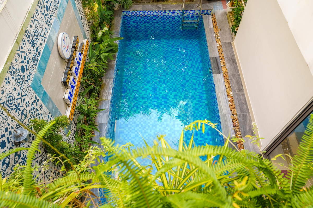 A well-kept swimming pool is surrounded by lush greenery and vibrant tiles, reflecting a tranquil blue hue. The area features a decorative tiled wall and is bordered by pebbles, offering a serene outdoor space for relaxation.