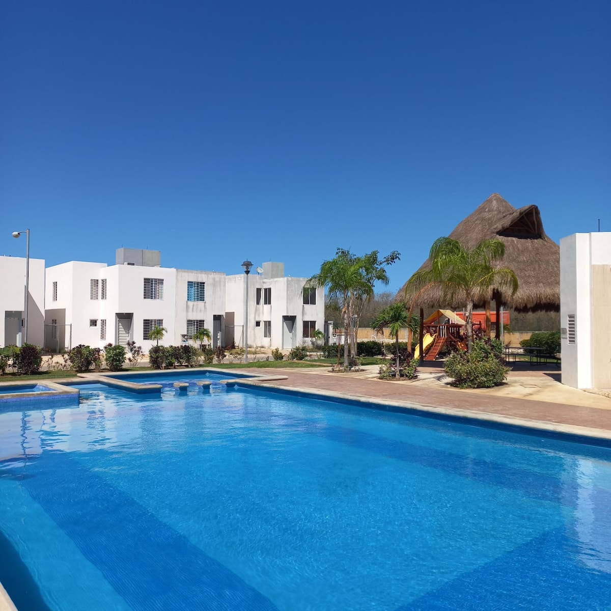 A clear view of the inviting swimming pool reflects the blue sky, surrounded by well-maintained gardens and modern white buildings. A palapa with a thatched roof is visible in the background, along with a playground area that includes colorful equipment.