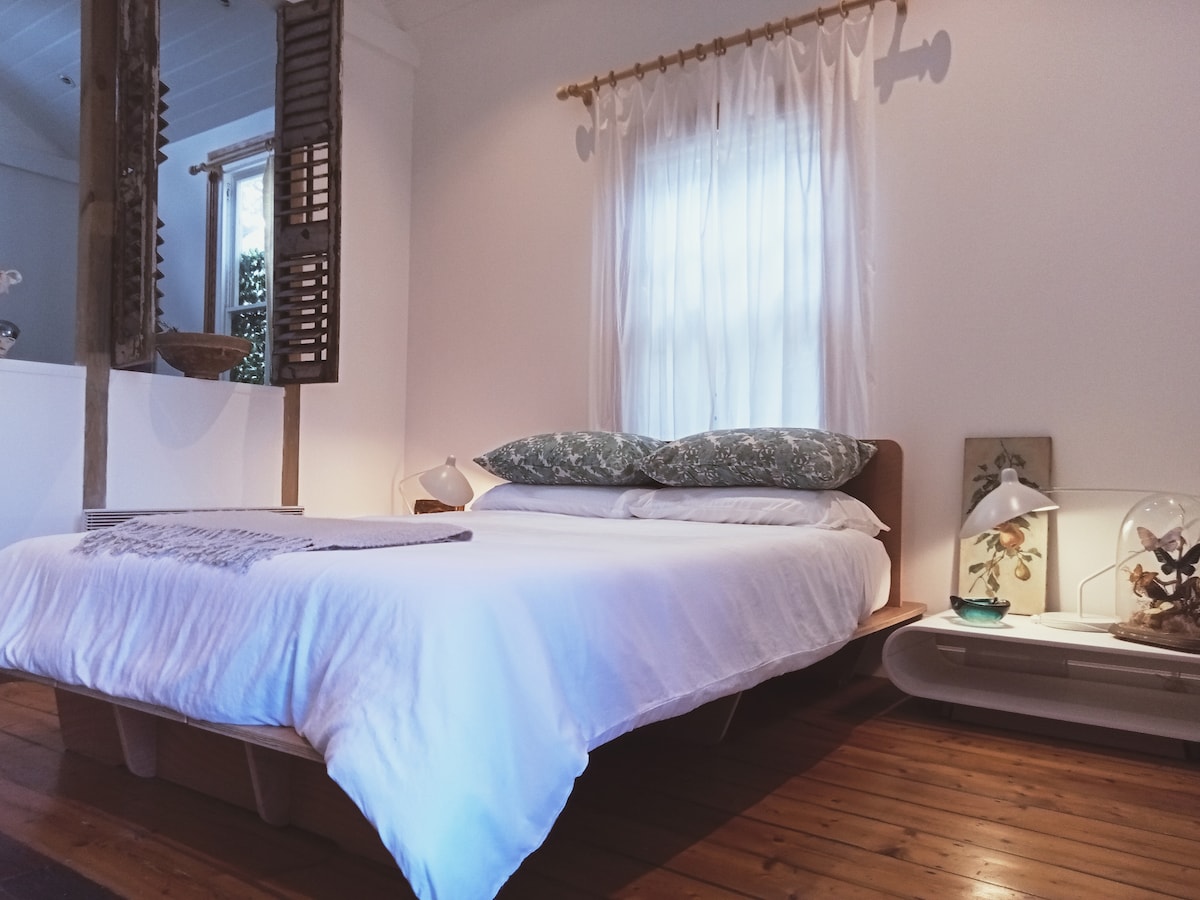 A cozy bedroom features a spacious bed with large, patterned pillows. Natural light filters through a sheer curtain, illuminating the warm wooden floor. Complementary bedside lamps are positioned on each side, and decorative artwork is displayed on the adjacent wall.