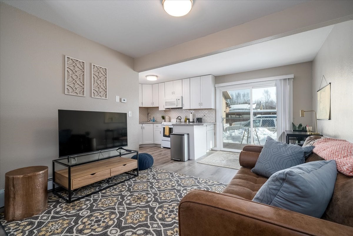 A modern living area is presented, featuring a comfortable sofa and a stylish area rug. A flat-screen television rests on a wooden media console. The open kitchen is equipped with sleek cabinetry and stainless steel appliances. Natural light fills the space from the sliding glass door leading to a patio.