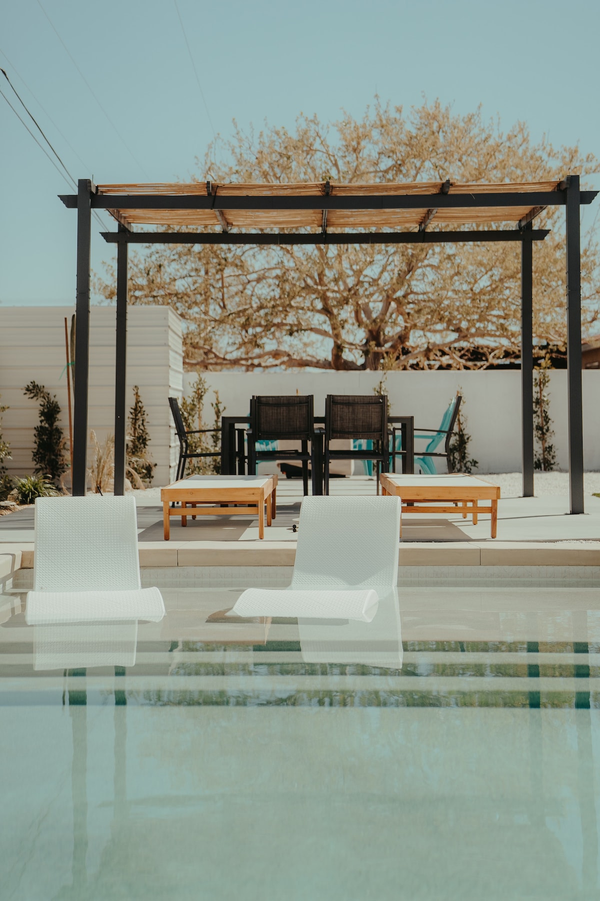 A serene lounging area is visible beside a non-heated pool, featuring two white in-pool loungers. A wooden pergola provides shade over additional seating, with a table and chairs situated under it. Lush greenery is seen in the background, enhancing the outdoor space.