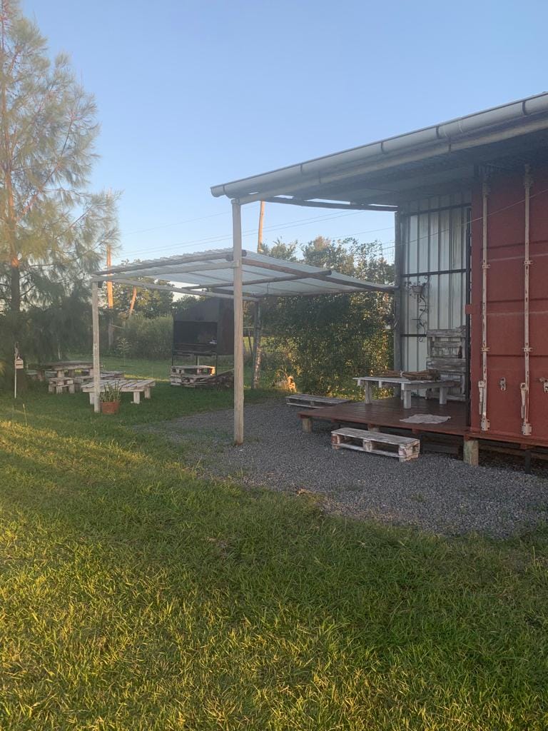 An outdoor area features a gravel surface with seating made from wooden pallets. A covered patio provides shade, while a container structure is visible in the background. Lush greenery surrounds the space, contributing to a calm environment.