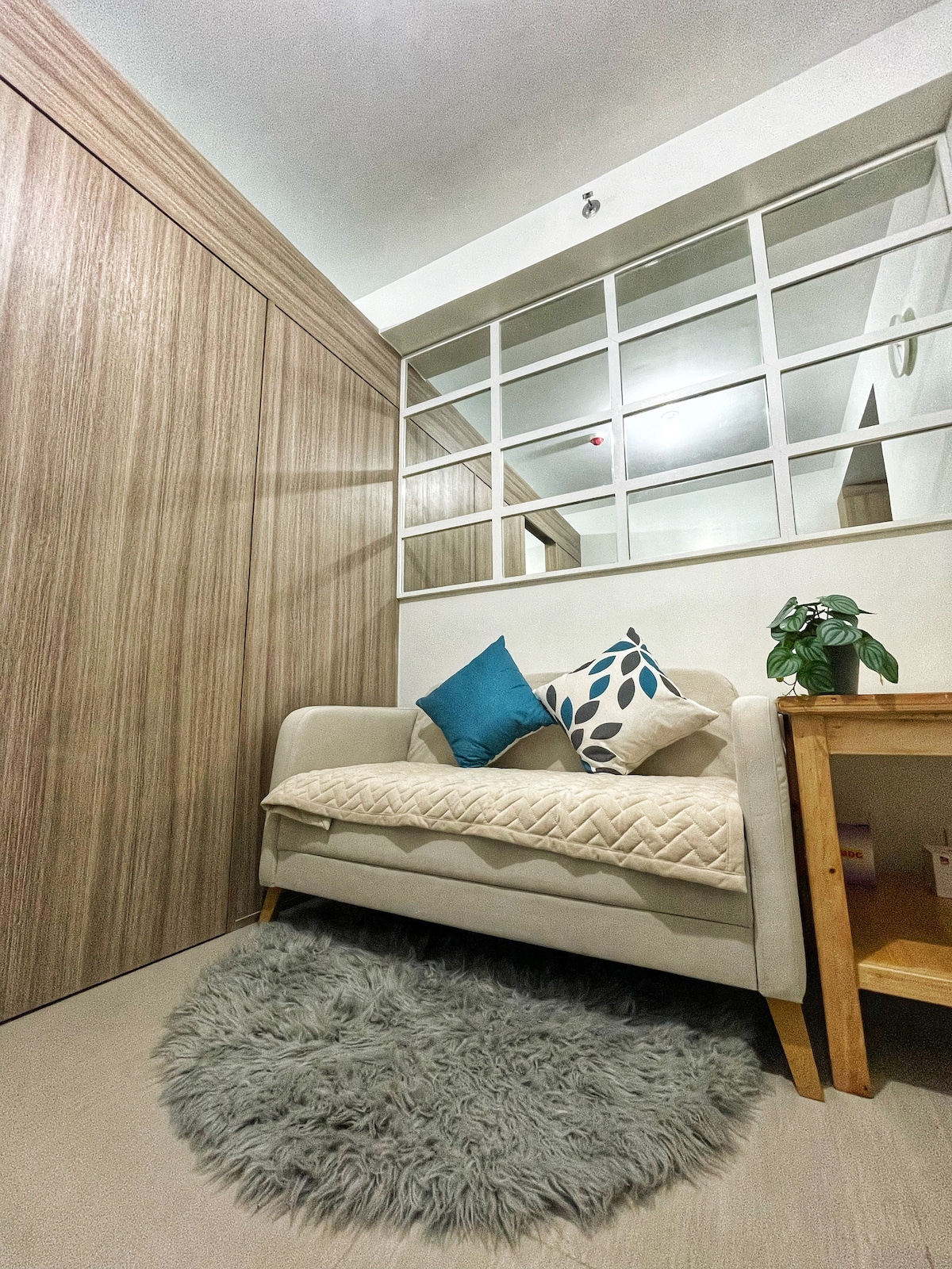 A cozy corner is presented with a light-colored sofa adorned with decorative pillows. A small wooden side table holds a potted plant. The wall features large glass panels that reflect the warm tones of the wooden cabinetry, enhancing the inviting atmosphere.