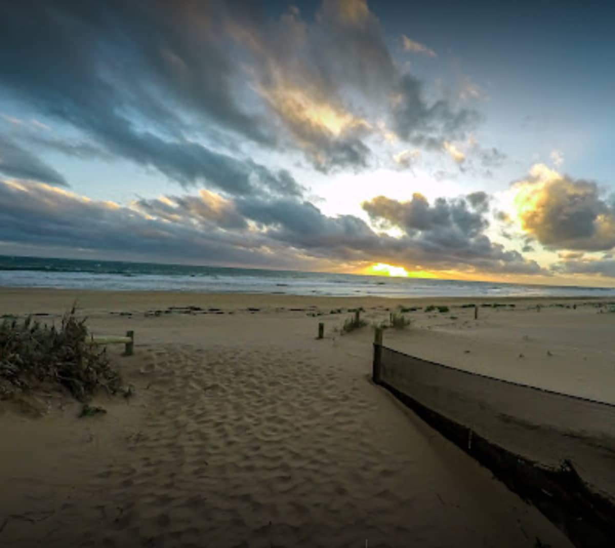 An expansive sandy beach is depicted, with gentle waves rolling in from the ocean. The horizon showcases a colorful sunset, highlighting hues of orange and yellow against a cloud-strewn sky. Beach grass and wooden posts are visible, marking the path leading toward the water.
