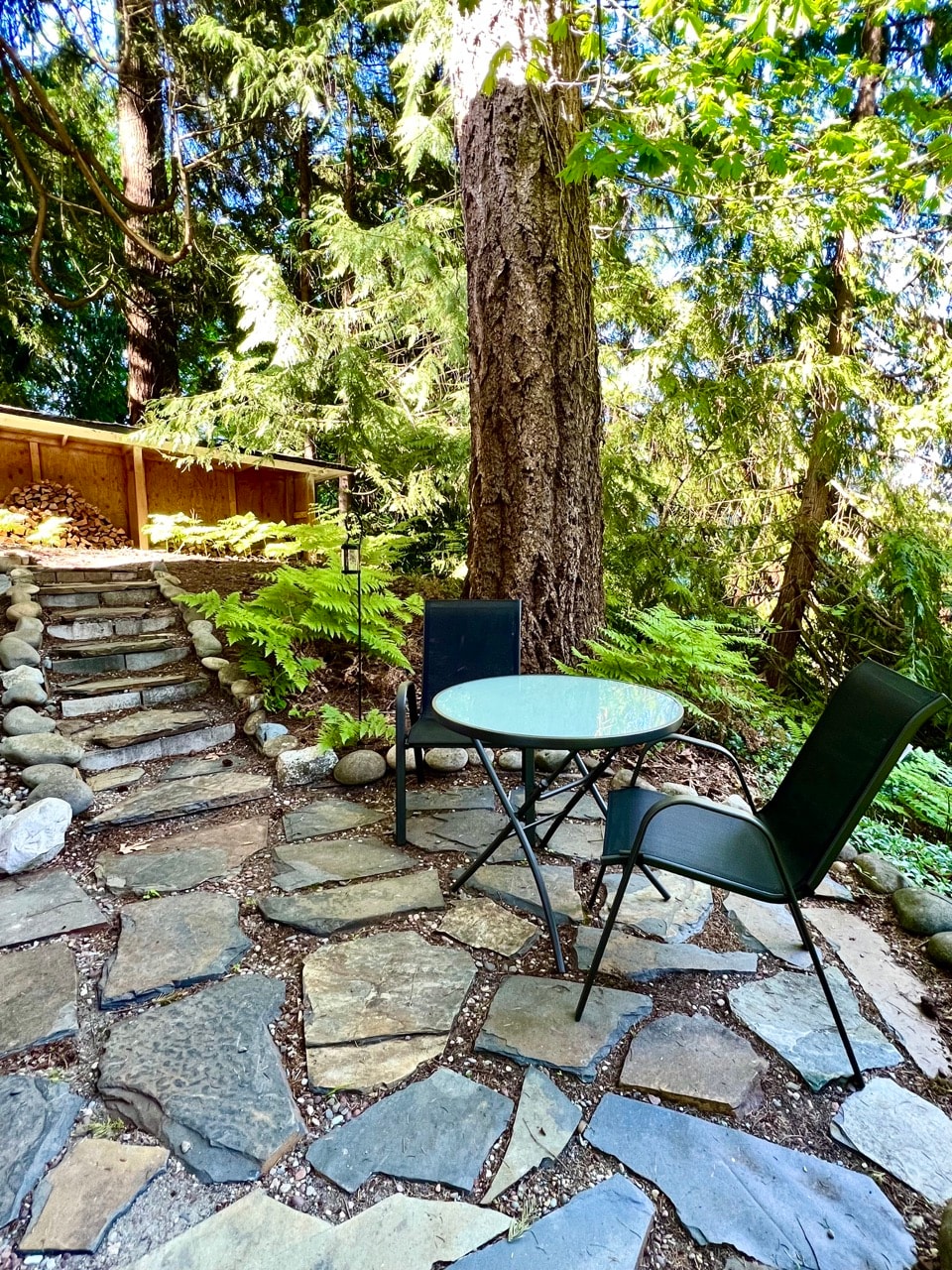 A natural stone pathway leads to a small outdoor seating area featuring a round glass tabletop and two black folding chairs. Lush greenery and large trees create a serene forest ambiance, complemented by a wooden structure visible in the background.