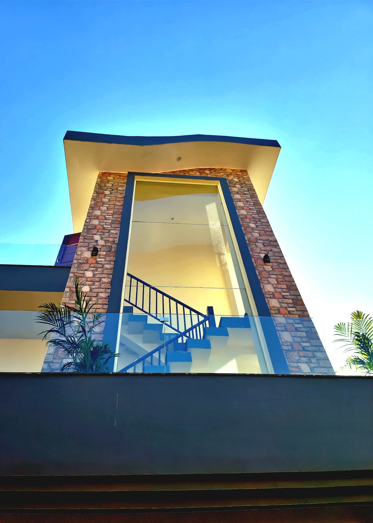 The exterior of the house showcases a striking design with a combination of stone and modern elements. A stairway leads up to the entrance, framed by large glass panels. The structure is set against a clear blue sky, enhanced by surrounding greenery.