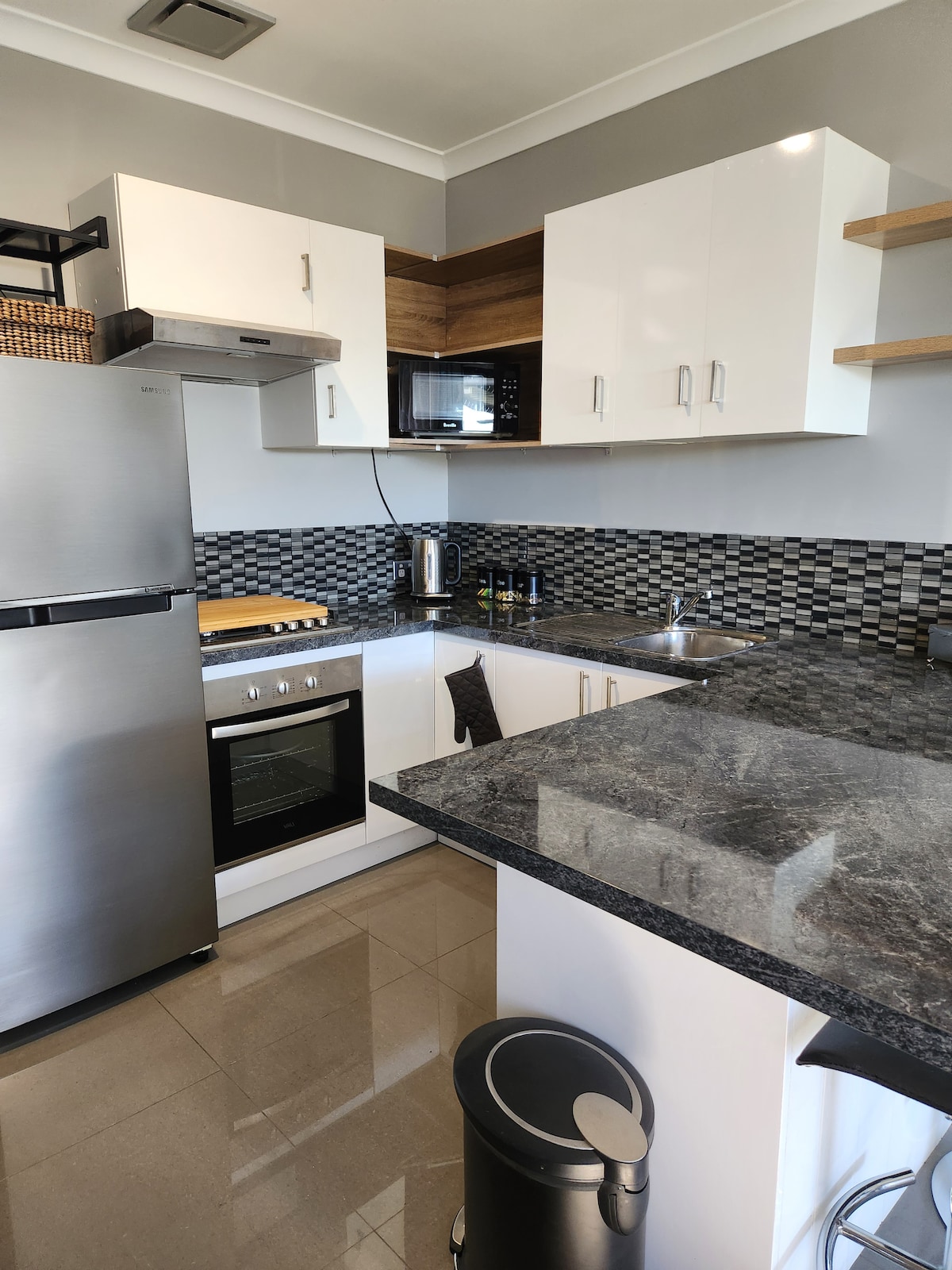 The kitchen features modern cabinetry with a combination of white and wooden accents. A stainless steel refrigerator stands next to a sleek oven and a black granite countertop, complemented by a mosaic tile backsplash. Essential appliances, including a microwave and kettle, are visible.
