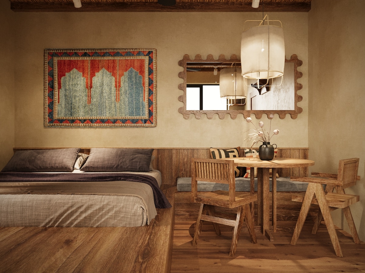 A warm interior showcases a spacious sleeping area with a large bed featuring neutral linens. A wooden dining table accompanies several wooden chairs. A decorative wall hanging and a mirror enhance the rustic design, complemented by soft pendant lighting.