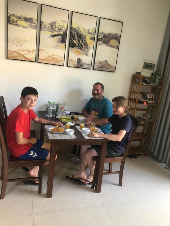 A dining area is shown with three individuals seated around a simple wooden table. Plates of food are arranged in front of them along with drinks. Artwork depicting a natural landscape hangs on the wall, and a shelf with books and decorative items is visible in the background.