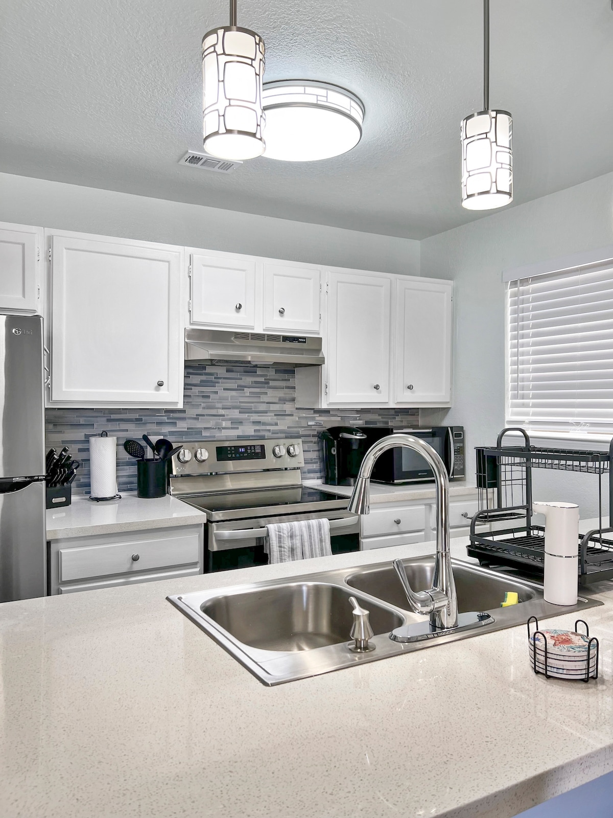 The fully equipped kitchen features white cabinetry and a modern design. Stainless steel appliances, including an electric stove and refrigerator, are present. A double sink sits beneath stylish pendant lighting, and a window provides natural light, enhancing the kitchen's functionality.