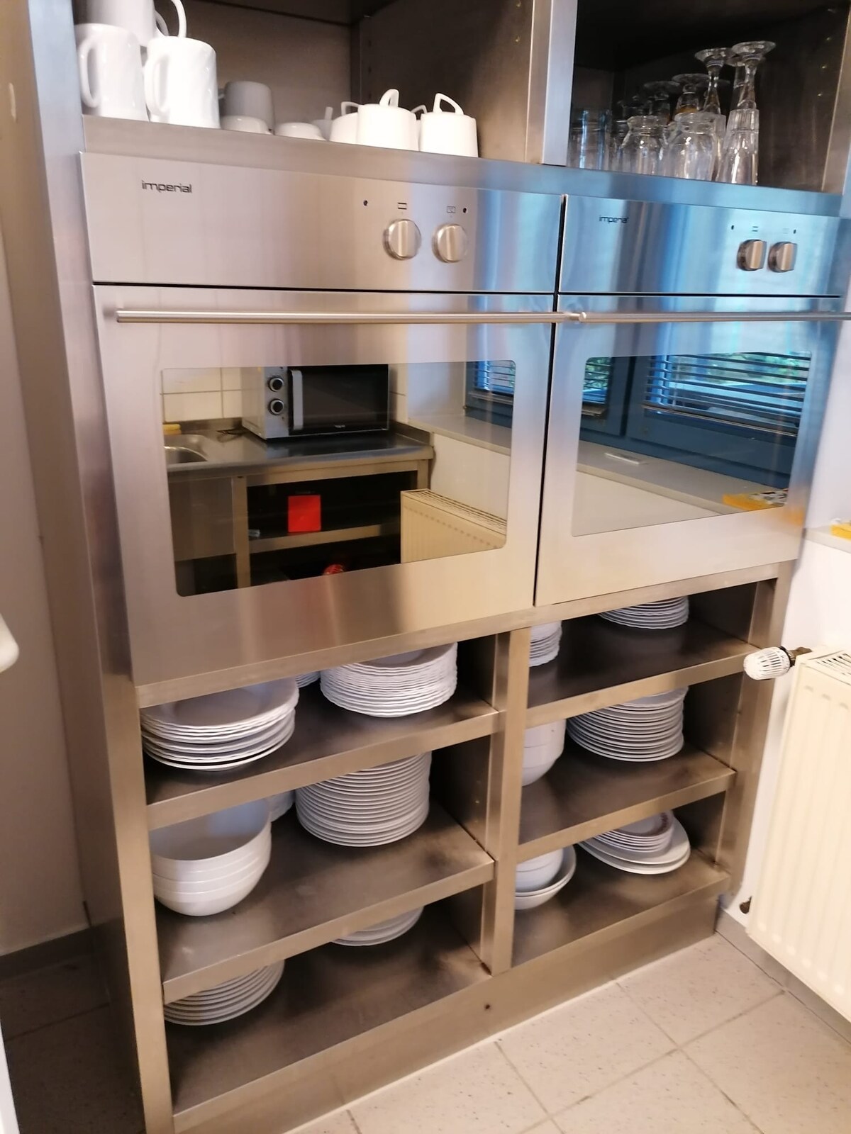 A metal kitchen cabinet showcases two built-in ovens with mirrored doors. Below, neatly arranged plates are stacked on shelves, and several glasses are visible above. A microwave is placed on a surface in the background, with natural light streaming through a nearby window.