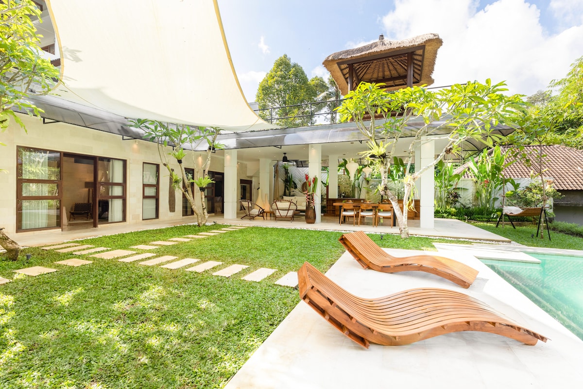A spacious outdoor area features a lush green lawn, two wooden lounge chairs, and a crystal-clear pool. A shaded deck area provides a dining space, while tropical plants enhance the serene environment. Large windows invite natural light into the villa's interior.