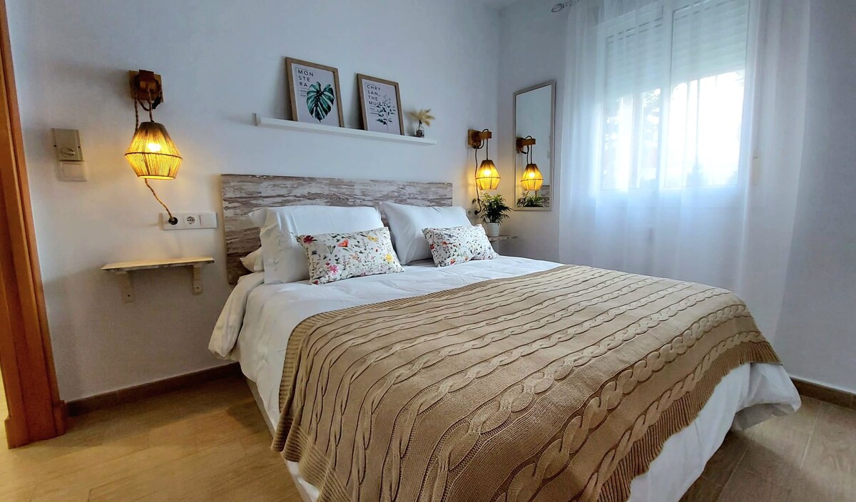 A spacious bedroom is featured, showcasing a large bed dressed in a textured beige blanket. Subtle floral pillows add a touch of color. Warm lighting is provided by wall sconces on either side, and natural light filters in through the window adorned with sheer curtains.