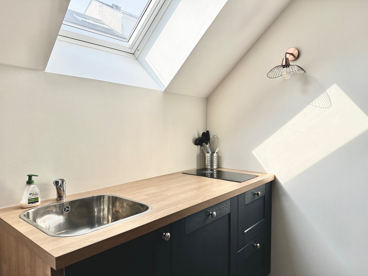 A modern kitchen area features a light wood countertop and dark cabinetry. A stainless steel sink is positioned beneath a skylight, providing natural light. Essential kitchen utensils are organized neatly nearby, complemented by a sleek light fixture that casts gentle shadows across the space.