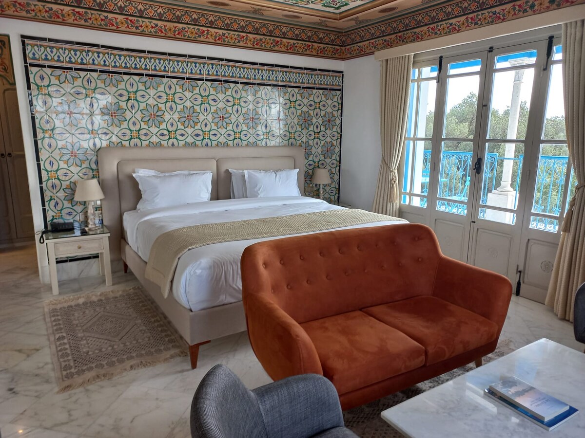 A spacious bedroom is highlighted by a patterned tile wall and features a comfortable bed with crisp white linens. A soft orange couch complements the room, while large windows provide natural light and views of the surrounding landscape. A stylish rug adds warmth to the marble floor.