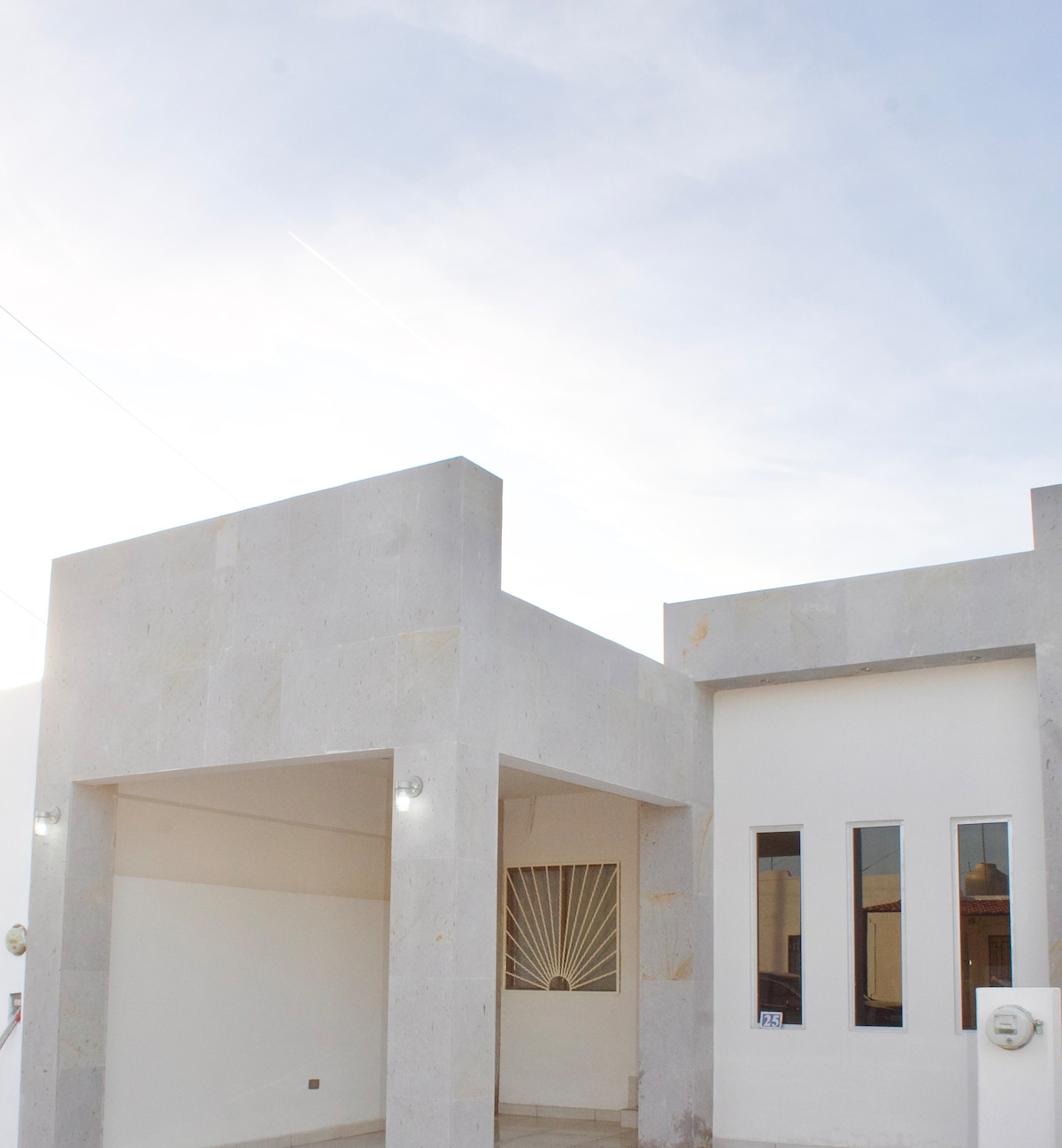 The exterior of the house features a minimalist design with light-colored stone walls. A small covered porch is seen at the entrance, flanked by large windows allowing natural light into the interior.