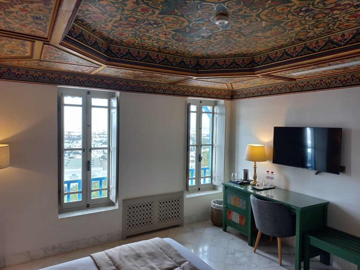 The room features ornate ceiling decor with intricate patterns. Large windows provide views of the city, allowing natural light to fill the space. A television is mounted on the wall across from a green desk with a lamp, while a comfortable chair is positioned nearby.