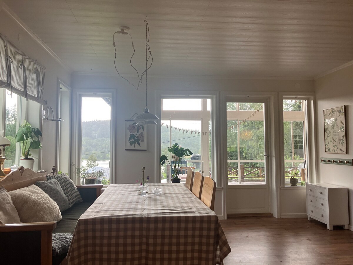A spacious dining area features a large table surrounded by wooden chairs, complemented by a cozy sofa. Bright windows offer views of the lake and greenery, while a decorative pendant light hangs above the table, enhancing the room's inviting atmosphere.