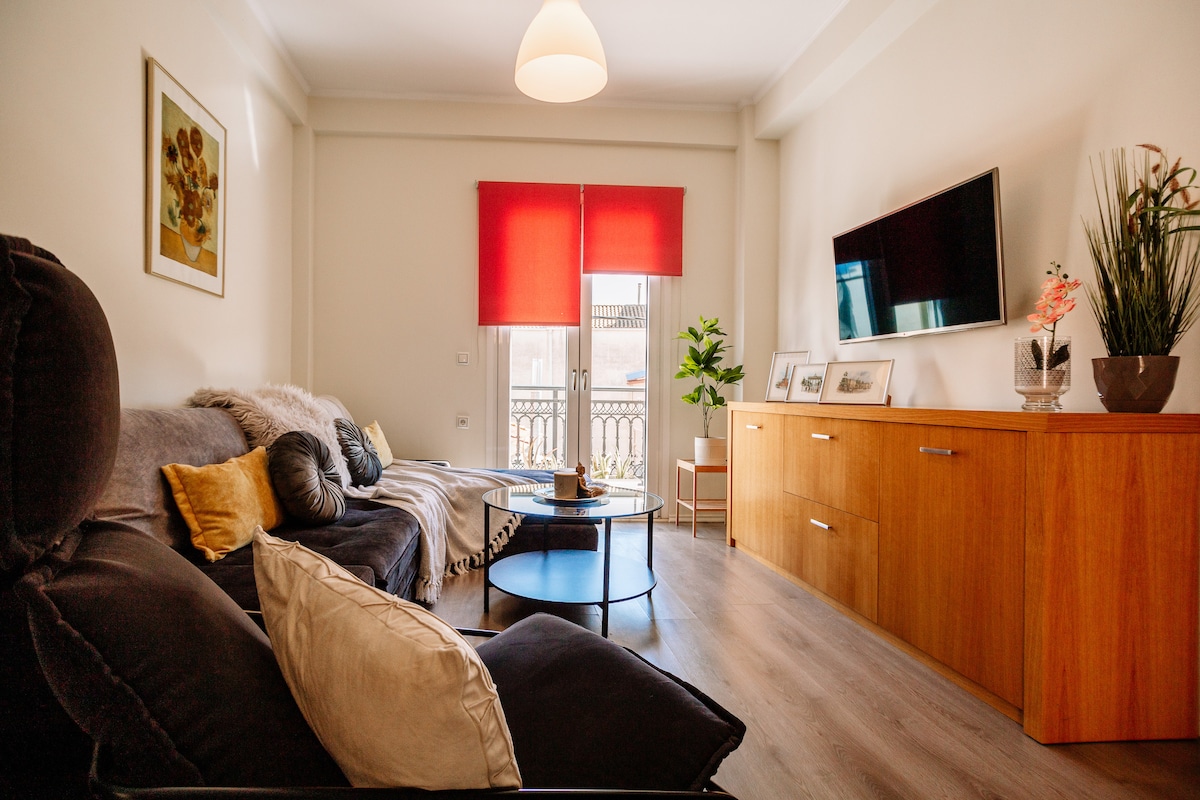 A bright living area features a dark fabric sofa adorned with decorative cushions. A wooden sideboard and a glass coffee table are positioned nearby. Large windows with red curtains allow natural light, while a television is mounted on the wall opposite.