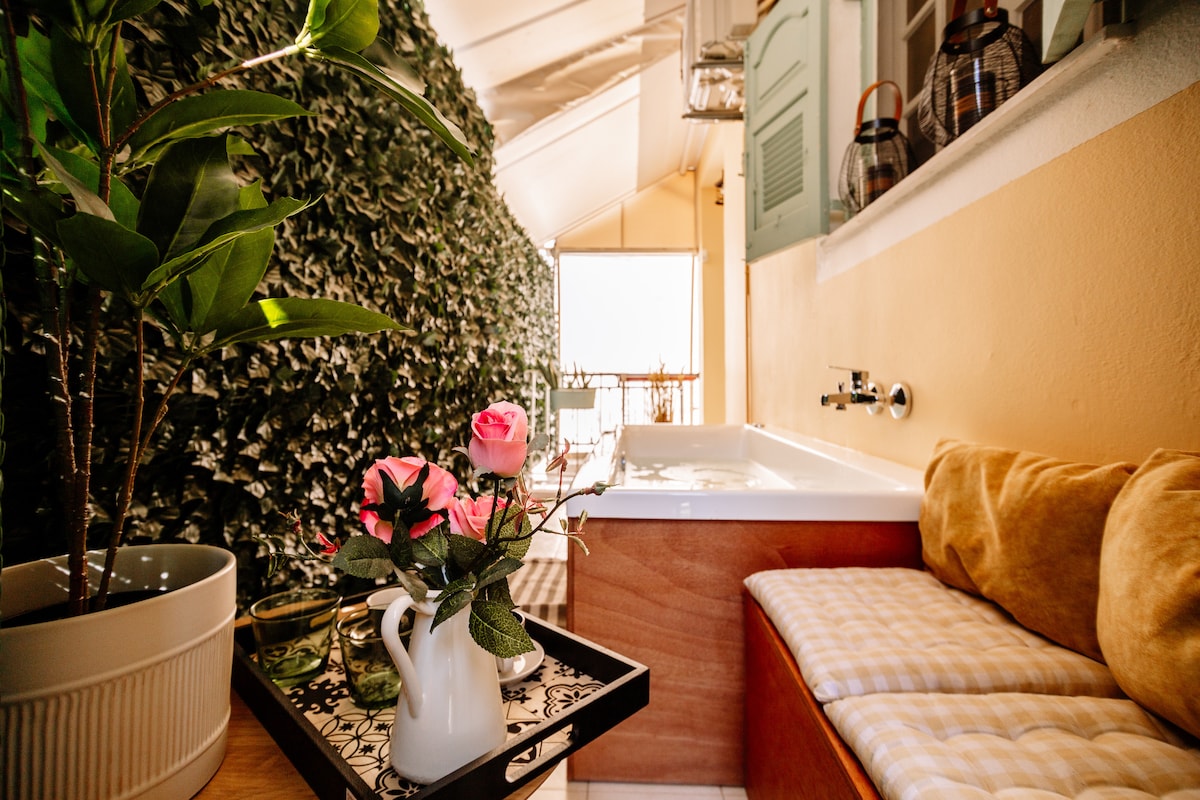 A cozy balcony space is highlighted by a hot tub and a small seating area. Fresh roses are placed on a tray beside a plant, while soft cushions provide comfort. Lush greenery adds privacy and a natural touch to the setting.
