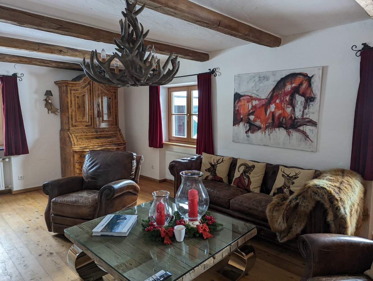 A spacious living area features wooden beams overhead and rustic furnishings. A comfortable sofa is adorned with decorative pillows, while an artistic horse painting hangs on the wall. A unique chandelier adds character, and a wooden cabinet enhances the warm ambiance.