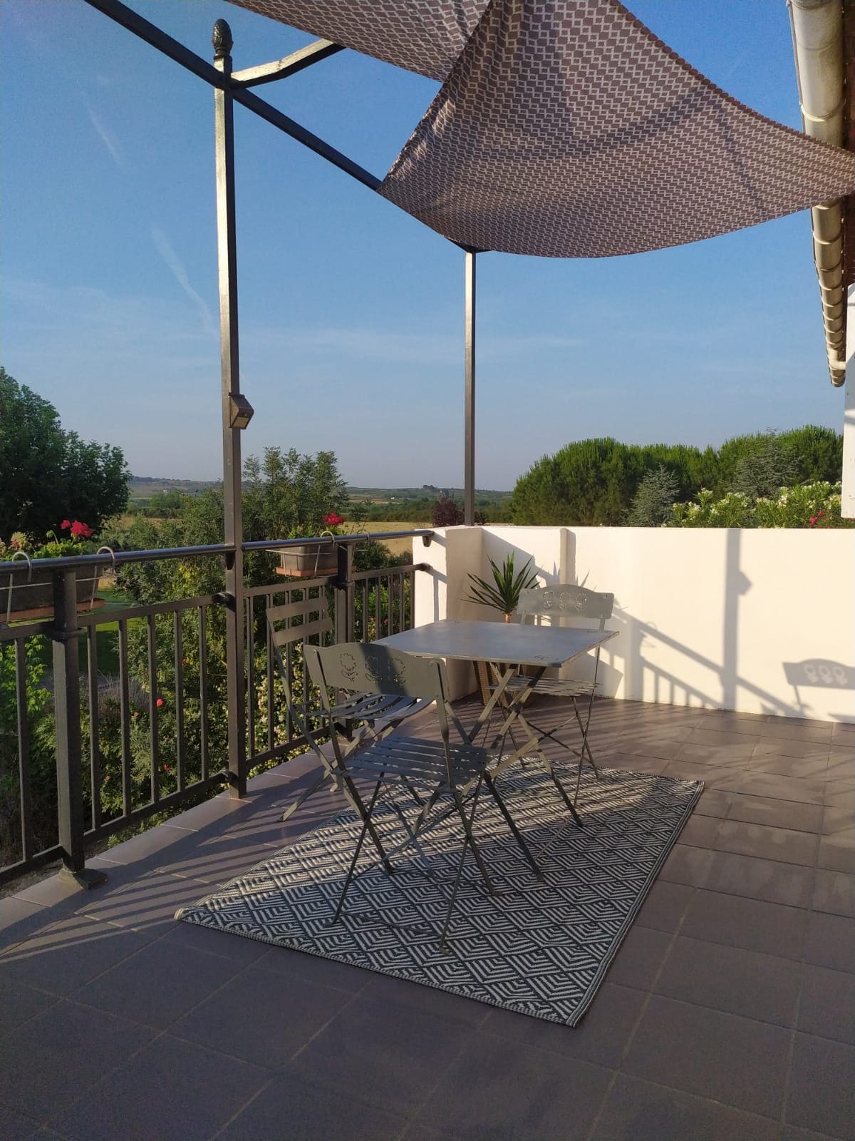 A shaded terrace features a round table and two metal chairs, surrounded by lush greenery. A geometric-patterned rug adds texture beneath the furniture. The railing offers an expansive view of the landscape, with a clear sky enhancing the outdoor ambiance.
