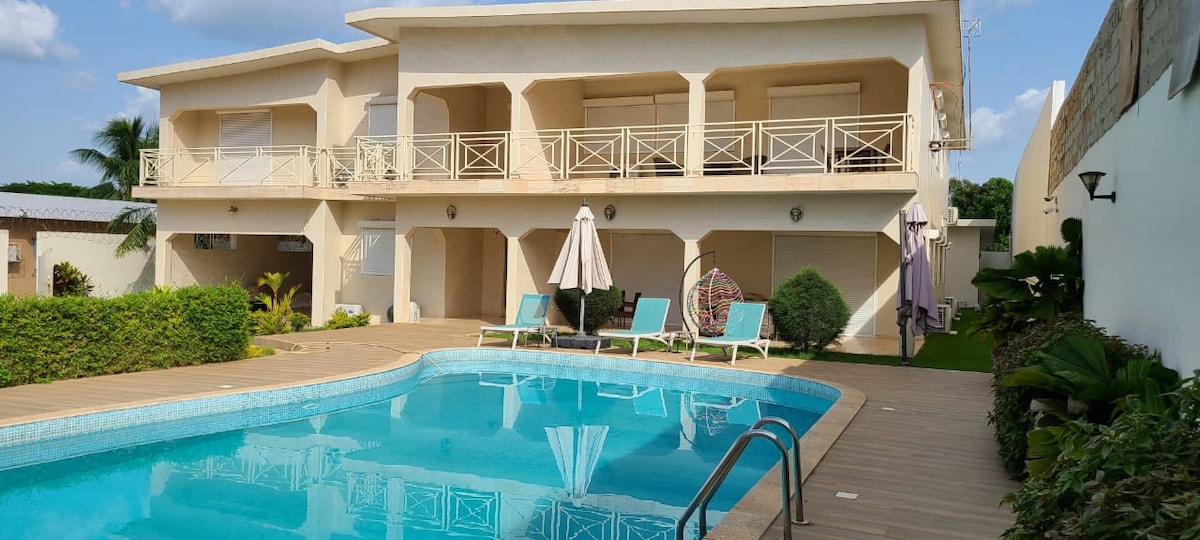 A large villa is shown with a private swimming pool. Sun loungers are positioned around the pool area, and a large patio is visible, offering ample outdoor space. The building features balconies with railings, surrounded by well-maintained greenery and palm trees.