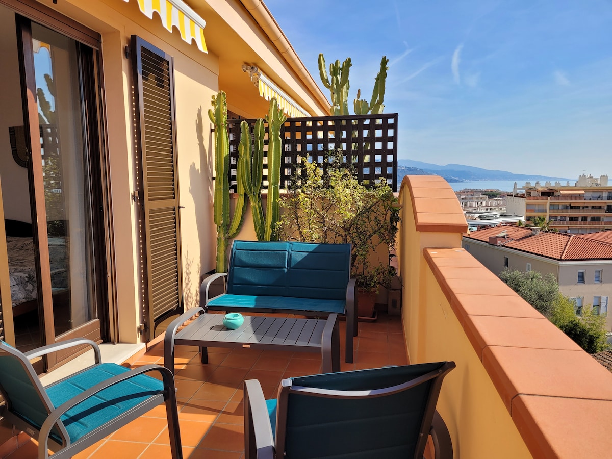 A spacious terrace features modern seating arrangements with two blue chairs and a gray table. Lattice screens provide privacy, and potted plants add greenery. The backdrop showcases distant views of hills and the sky, enhancing the overall outdoor ambiance.