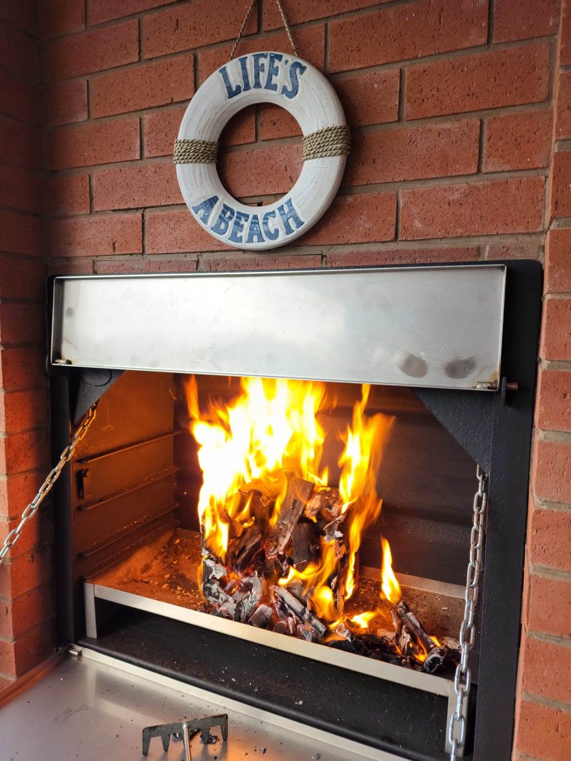 A built-in braai is showcased against a backdrop of red brick, with flames roaring optimally. Above, a decorative wooden lifebuoy featuring the phrase 'LIFE'S A BEACH' is hung on the wall, adding a coastal charm to the space.