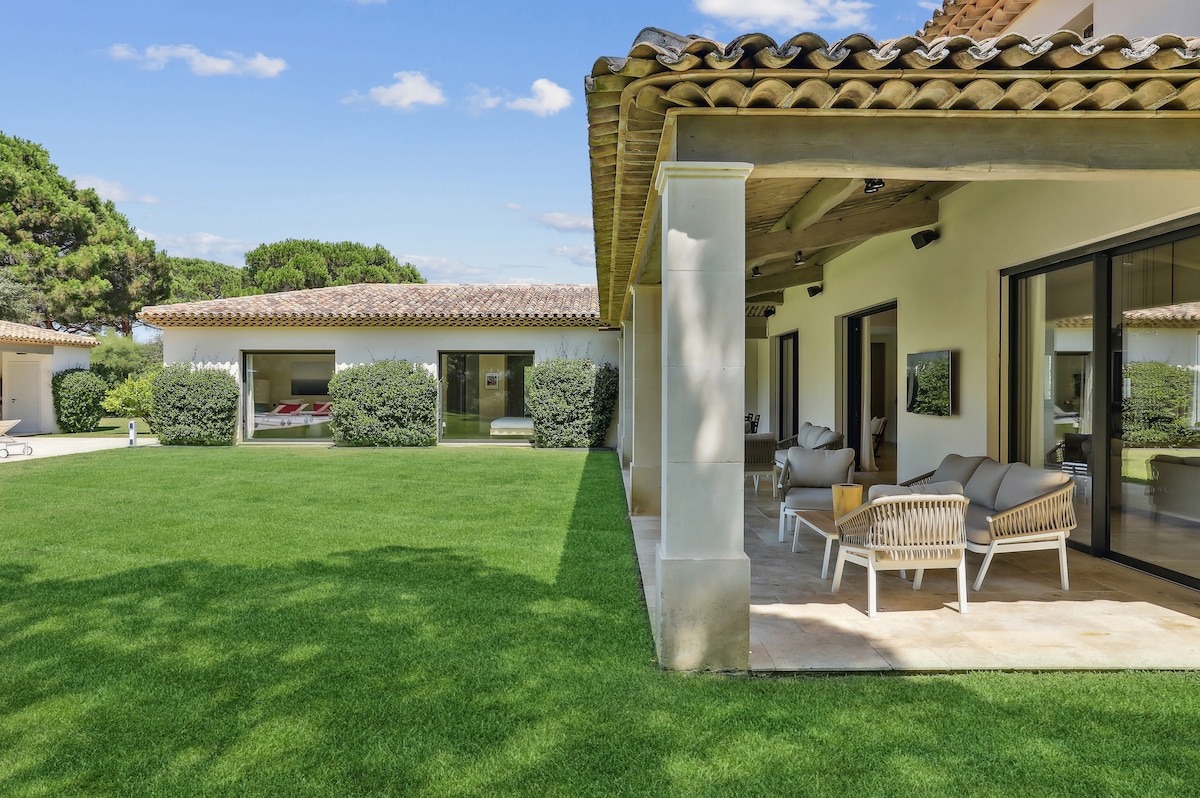 A spacious outdoor area features a well-manicured lawn, bordered by low greenery. Covered seating with light-colored cushions is positioned on the terrace, providing a comfortable space to relax. The villa's exterior showcases a tiled roof and large glass doors leading to the interior.