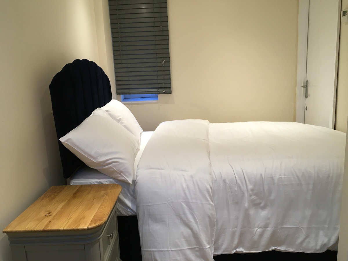 A cozy bedroom features a spacious bed adorned with white linens, complemented by a wooden nightstand. Natural light enters through a window with closed blinds, creating a bright yet private atmosphere. The room is neatly arranged and offers a calming space for rest.