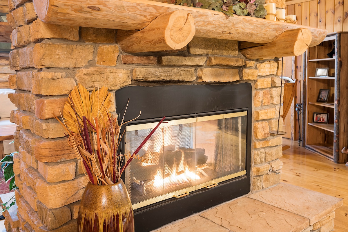 A stone fireplace is centered within a wooden interior, featuring a clear glass front that displays the flickering flames. Above, a rustic wooden mantel holds decorative elements, while a ceramic vase with dried grasses is placed beside the fireplace.